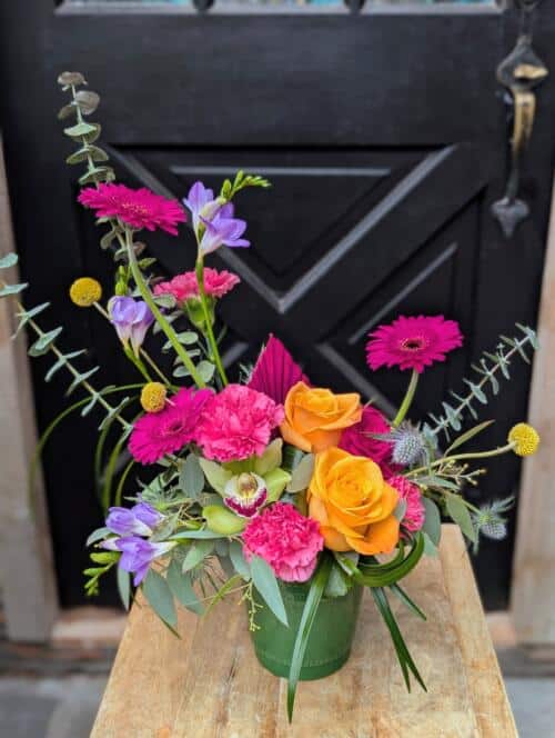 Front view of a green vase with orange roses, pink gerberas and carnations, purple freesia and eucalyptus, by a black door.