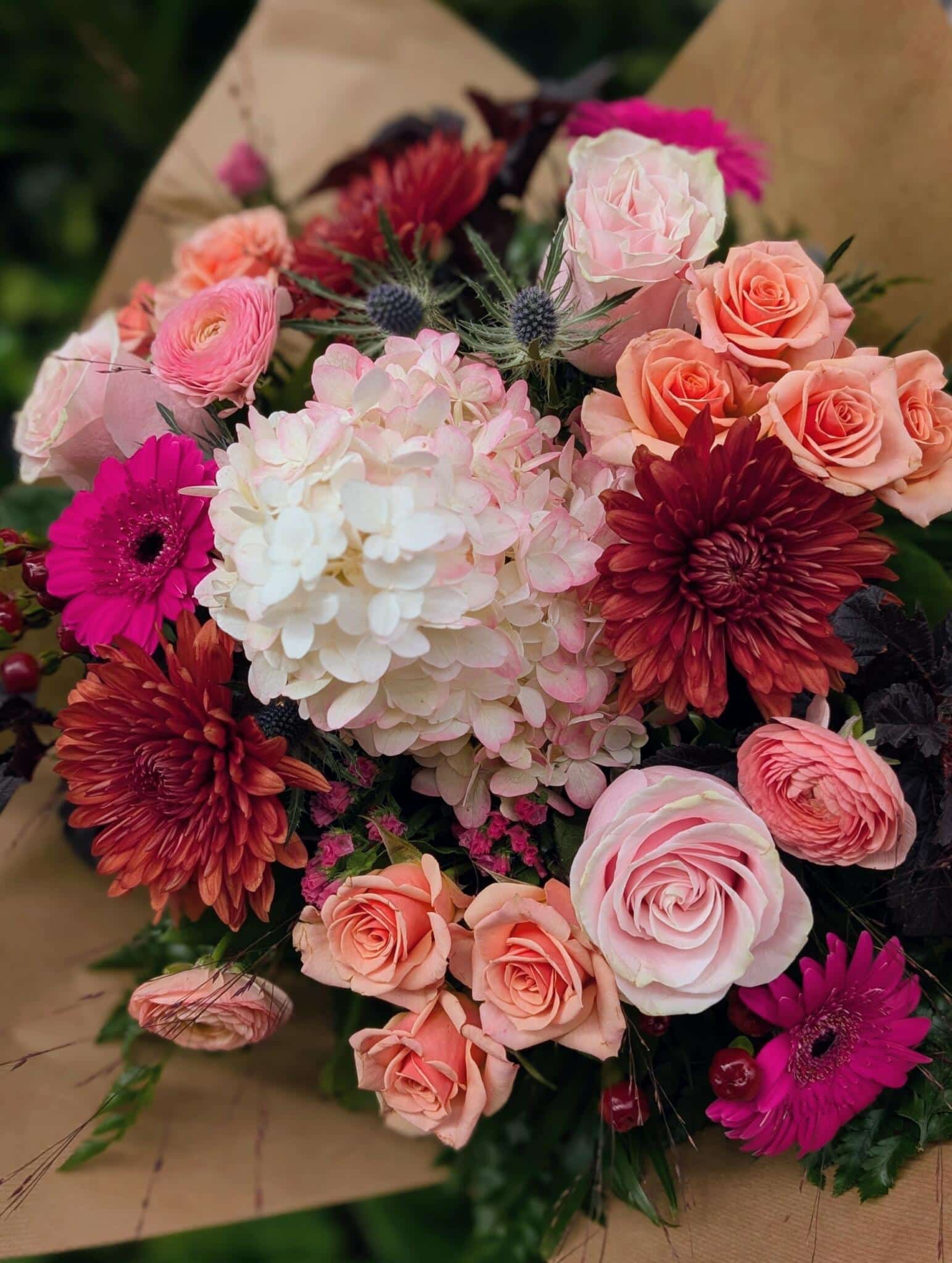 Close view of a bouquet of peach roses, pink gerberas, red dahlias, a white hydrangea and blue thistle in kraft paper.