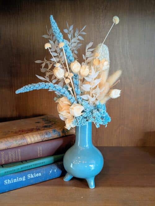 Dried arrangement of blue-dyed stems and fillers with peach and cream blooms in a teal footed vase, beside old books.