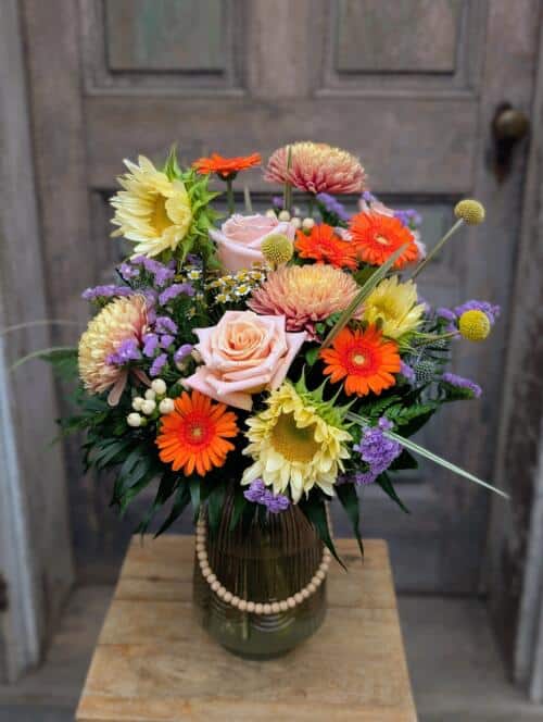 Front view of a beaded glass vase with peach roses, orange gerberas, sunflowers, dahlias and purple statice by a grey door.