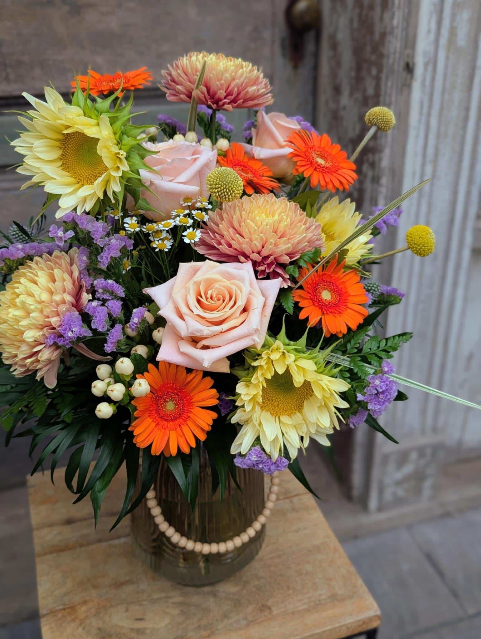 Angled view of a vase arrangement with a peach rose, orange gerberas, yellow sunflowers, peach dahlias and purple statice.