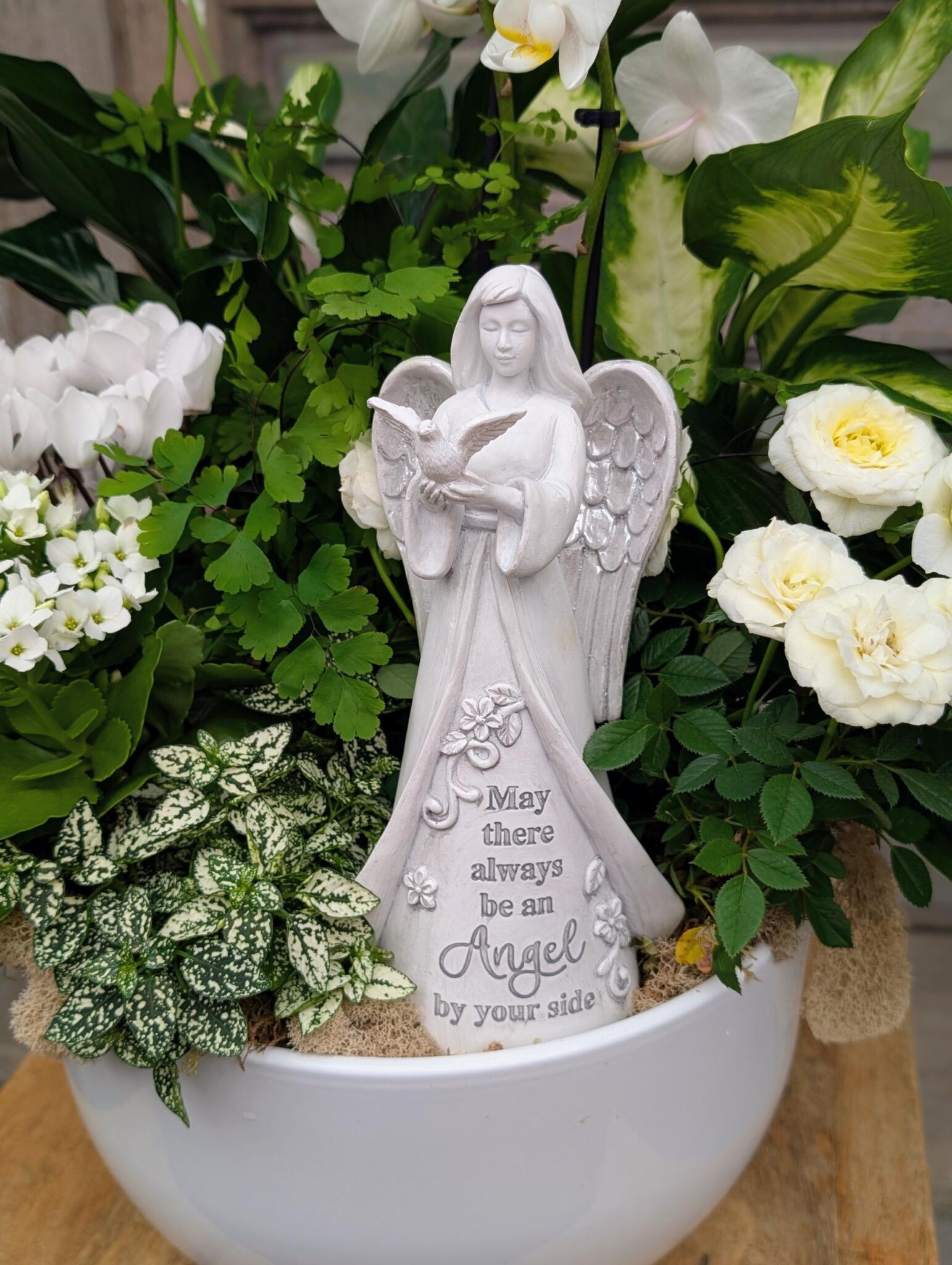 Close-up of a white angel figurine reading “May there always be an Angel by your side” among white blooms and variegated foliage.