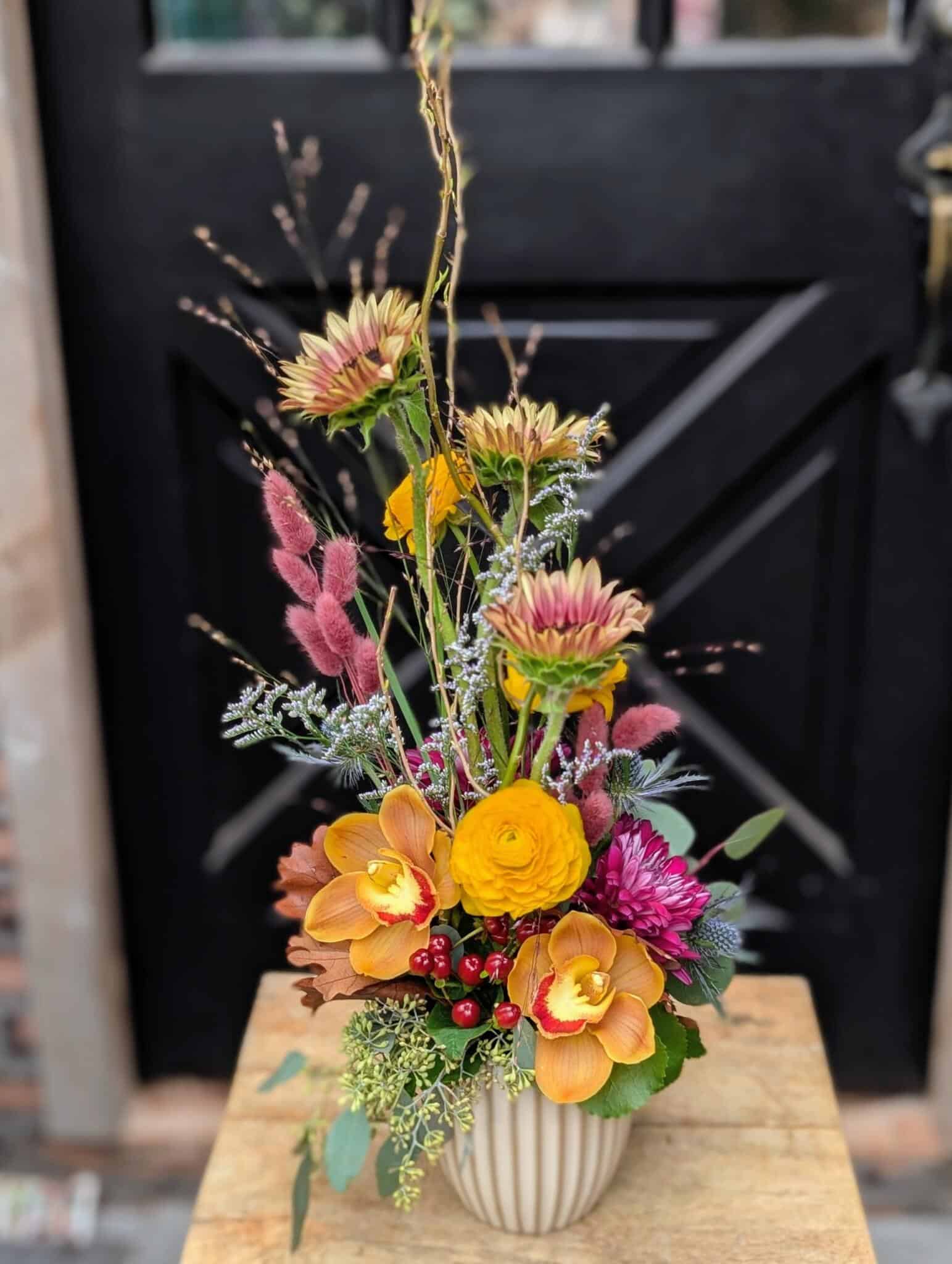 Tall arrangement of orange orchids, a yellow bloom, magenta mum and pink plumes in a ribbed cream vase by a black door.