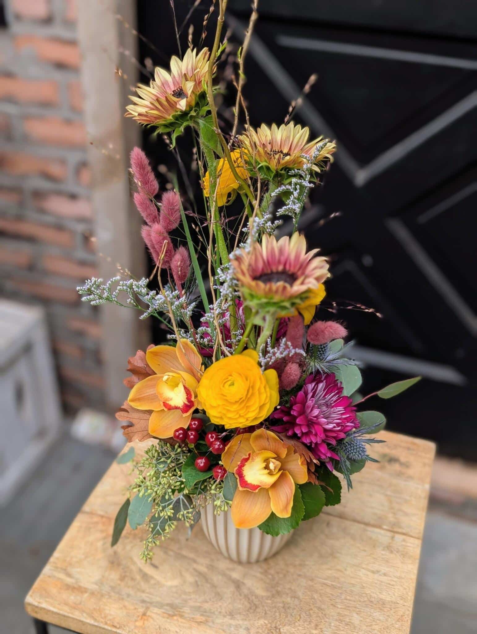 Angled view of orange orchids, a yellow ranunculus, a magenta mum and pink plumes in a tall arrangement in a cream vase.