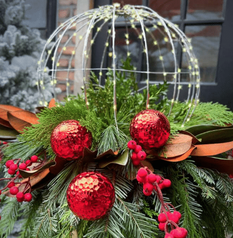 Winter planter of evergreen boughs, red ornaments, red berries and magnolia leaves under a lit wire sphere.
