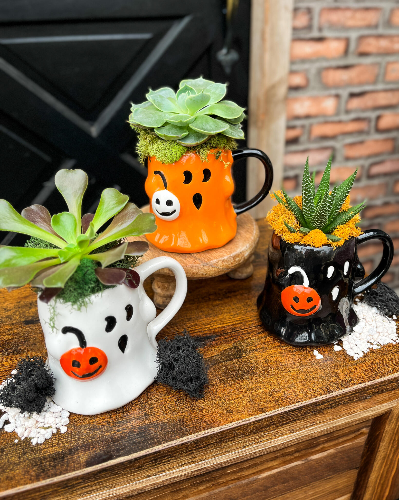 White, orange and black ghost mug planters with succulents, set at different heights beside a brick wall.