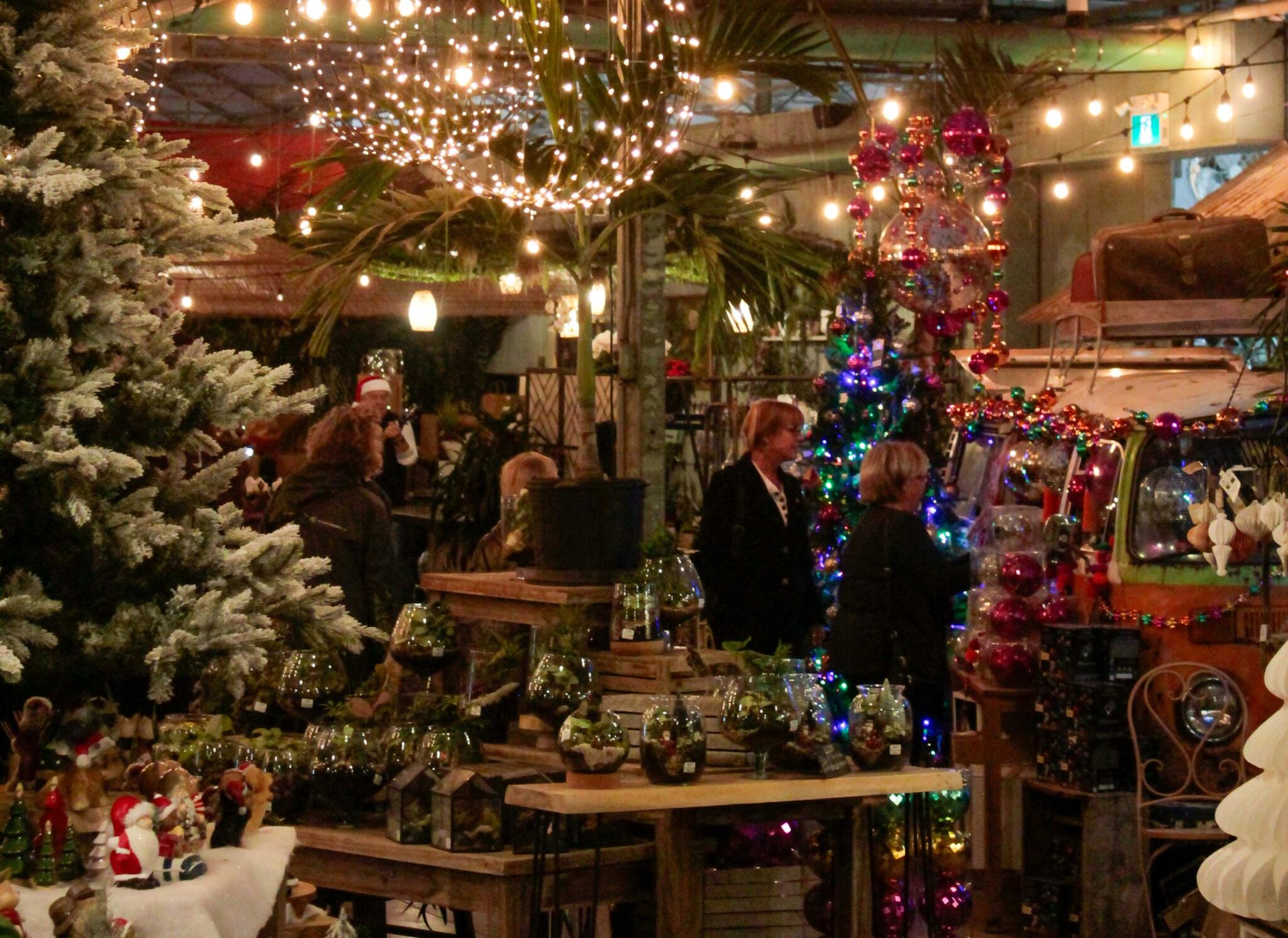 Late-Night Browsing in the Holiday Greenhouse Shoppers browse holiday displays in a greenhouse lit with string lights, frosted trees and ornaments.