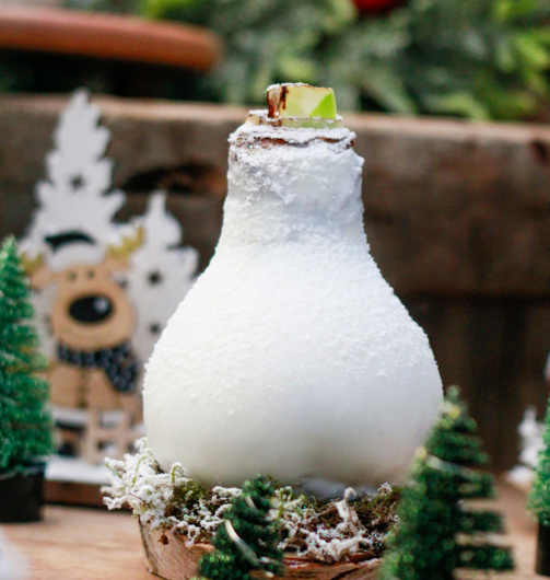 A white wax-dipped amaryllis bulb nestled in moss with tiny evergreen trees and festive figurines.