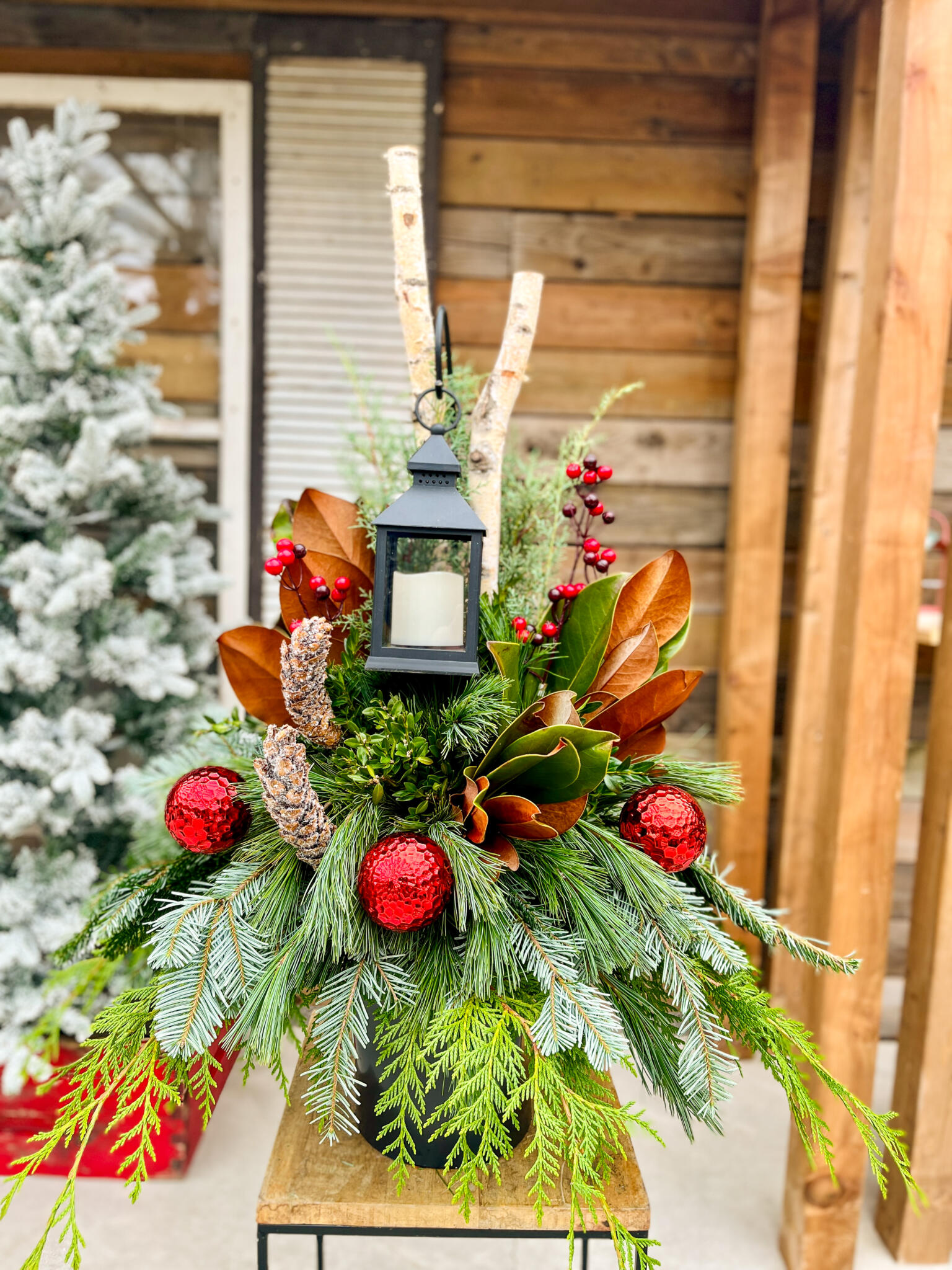Outdoor winter urn with a black lantern, birch poles, magnolia leaves, red ornaments, pine cones and mixed evergreens.