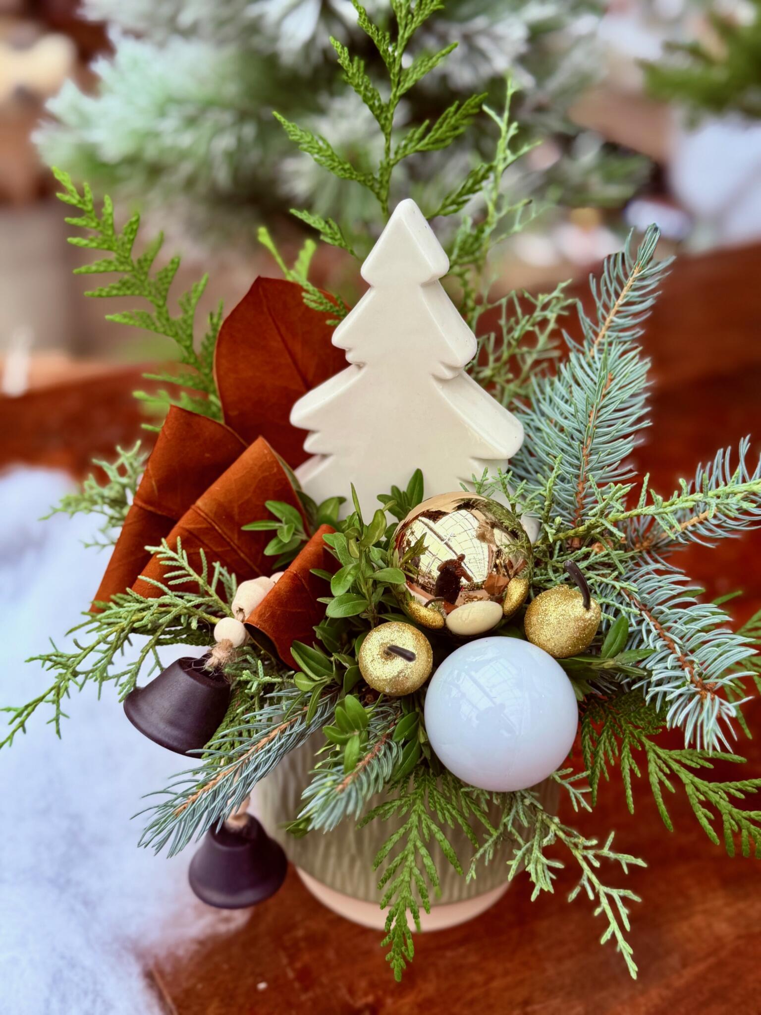 Small winter arrangement with a white ceramic tree, gold and white ornaments, dark bells, rust leaves and evergreens.