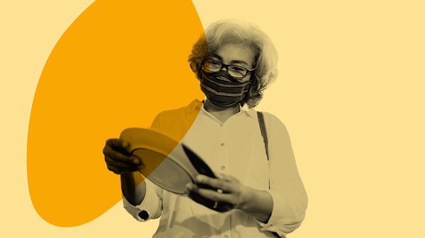 A masked woman dressed in a white, button-down blouse examines a dinner plate.