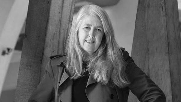 British professor Mary Beard pictured in black and white, from the chest up. She has light skin, light hair that falls below her shoulders, wears a black shirt and trench coat, and is smiling.