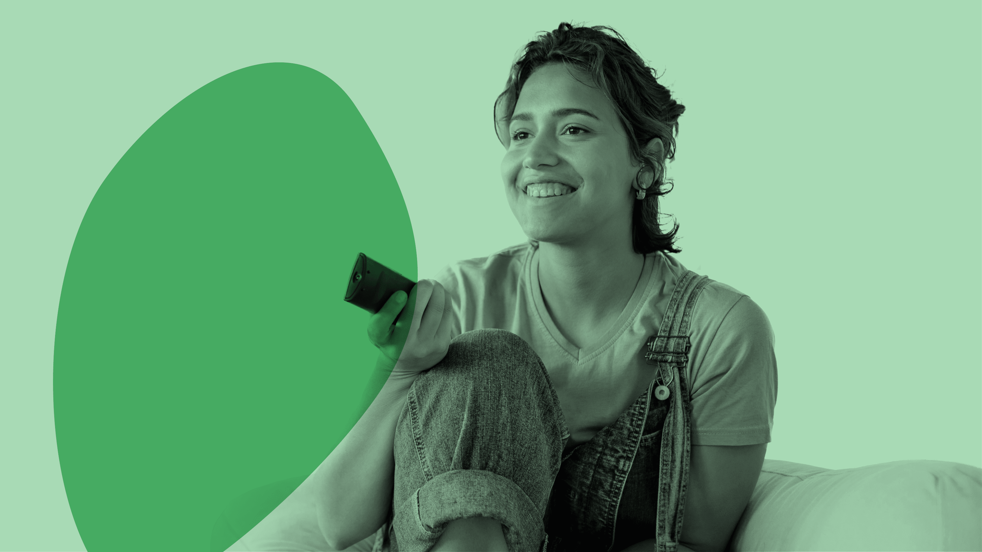 A woman dressed in overalls and a T-shirt sits on a couch smiling while pointing a TV remote.