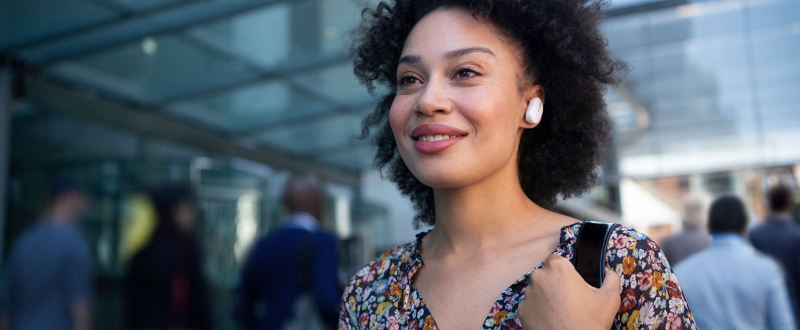 A woman in a floral-themed top is pictured in a busy walkway. A white earbud is visible in her left ear.