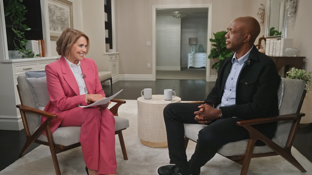 Future Ready: Artificial Intelligence with Katie Couric and Google SVP James Manyika. Couric has light brown hair, light skin, and wears a coral pantsuit. Manyika has a shaved head, dark skin, and wears a black jacket, collared shirt, and black pants.
