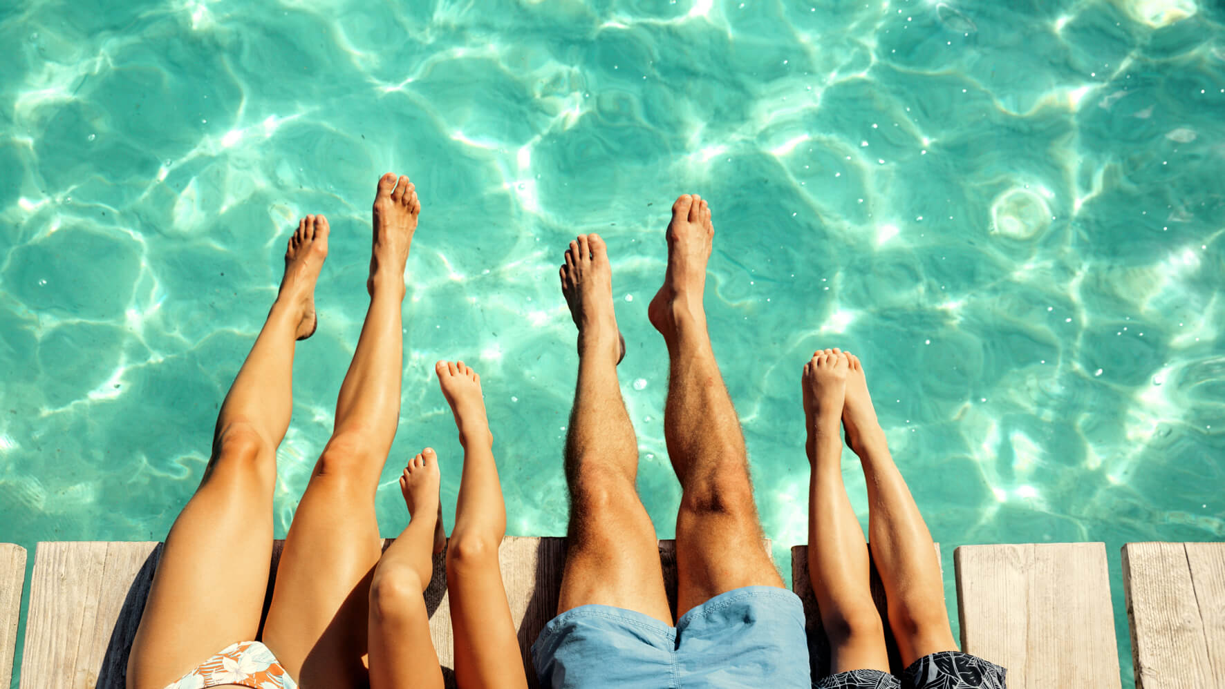 A family of four sit at the side of a swimming pool with their feet dipped into the water.
