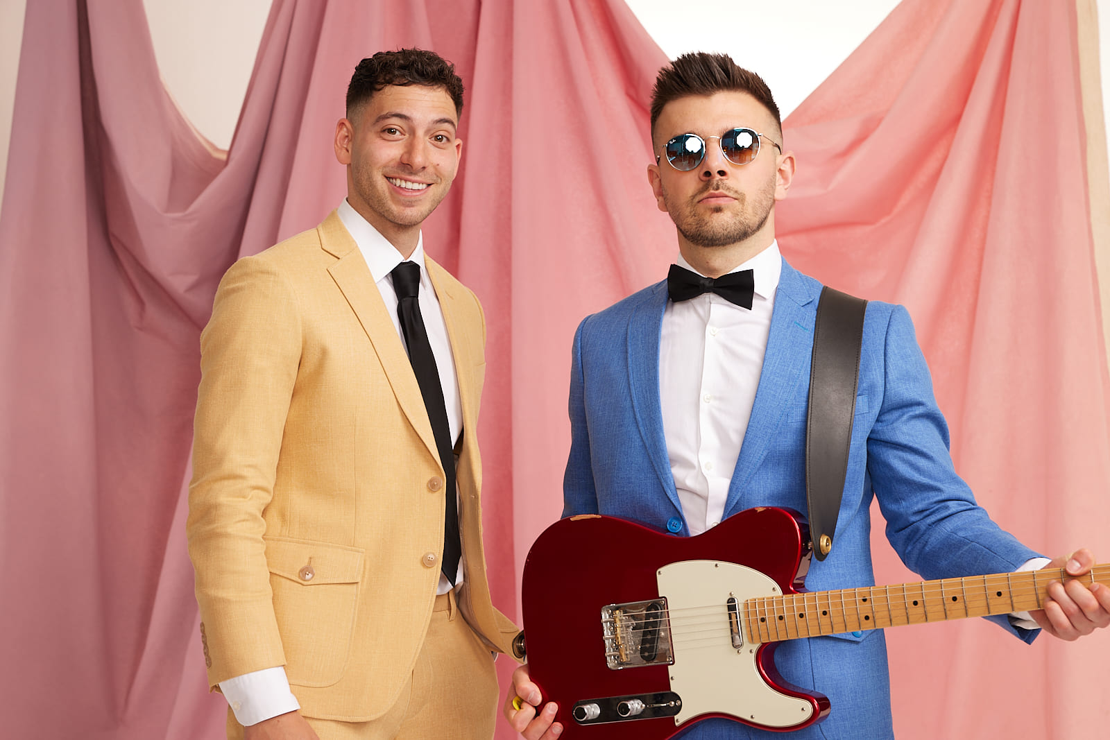 Music duo Crash Adams is pictured wearing suits. The man on the right also wears sunglasses and holds an electric guitar.