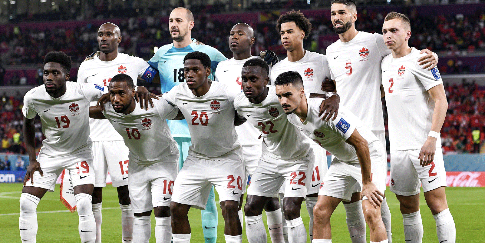 The Canadian men’s World Cup soccer team, assembled in two rows and wearing white jerseys, shorts, and socks, poses as a group at the 2022 FIFA World Cup.