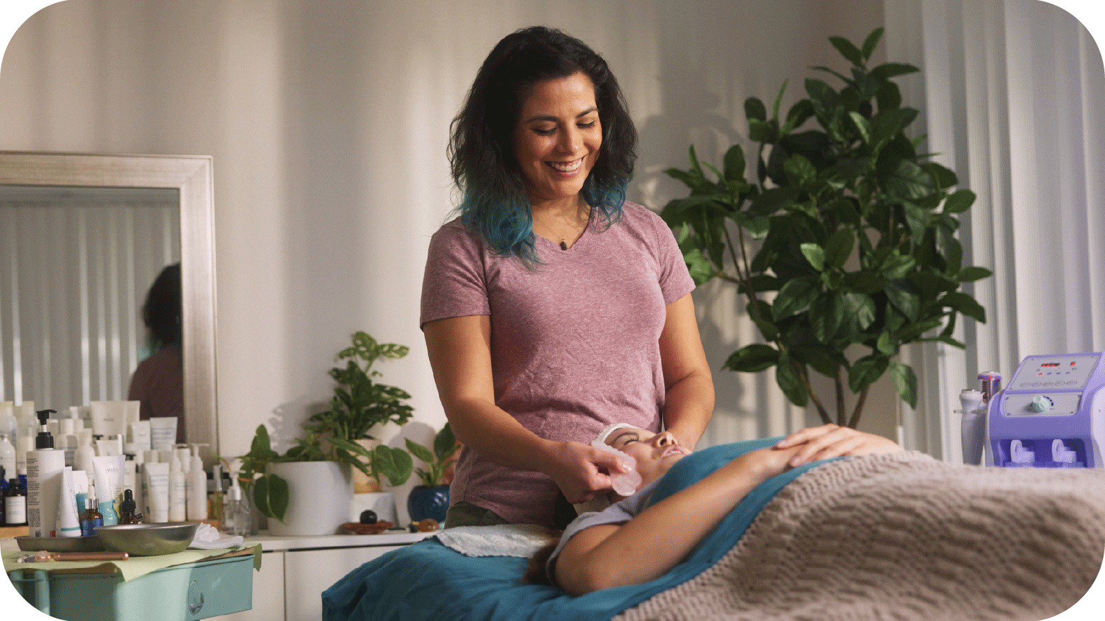 A smiling Lety Soto, pictured in casual clothing with shoulder-length hair that’s dyed light blue on the ends, stands over a client resting on a massage table and applies a facial technique aimed at relaxation and stress relief.