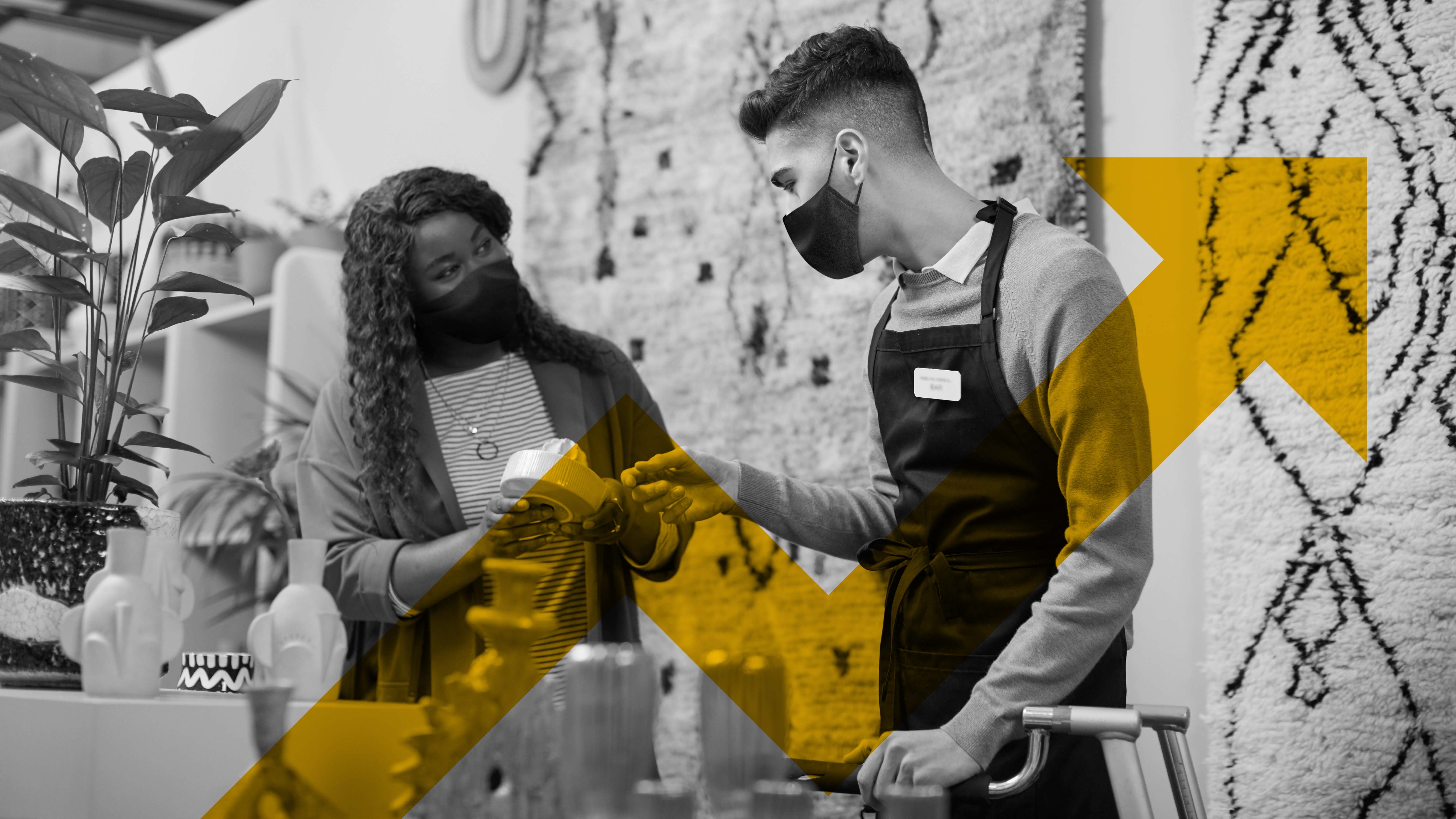 A black-and-white photo of two individuals inside a store: a woman wearing a face mask speaks with  a male employee in a face mask and shop apron. Overlaid on top of the image is a stylised translucent yellow tick pointing upwards.