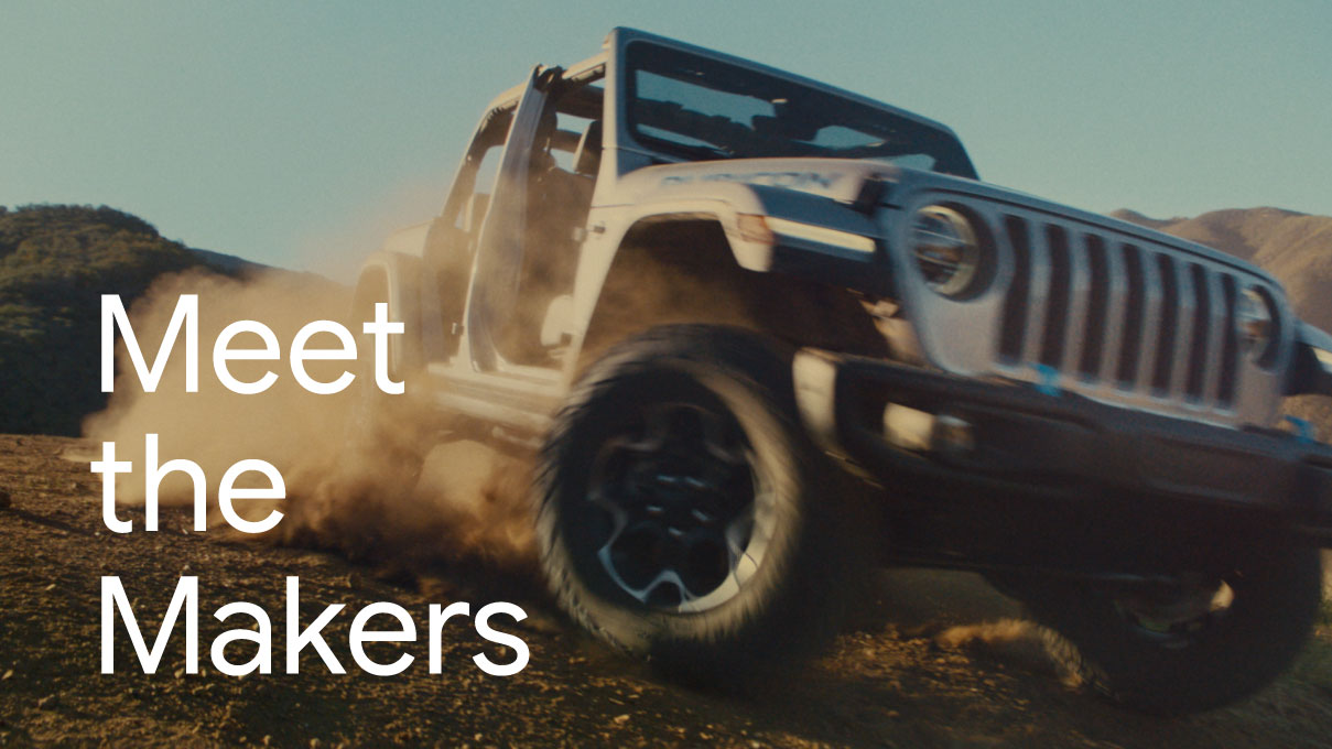 Meet the Makers: A Jeep Wrangler 4xe kicks up a cloud of red dirt while making a sharp turn on a rocky desert knoll.