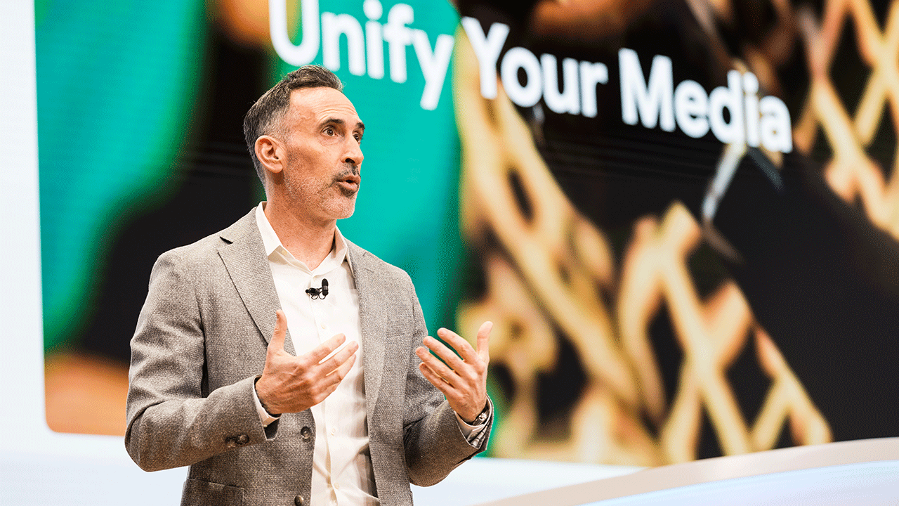 Sean Downey, president, Americas & Global Partners at Google, speaks to the audience at 2024 IAB NewFronts in front of a screen that reads “Unify Your Media” in white lettering on a gold and green image that includes a hand holding a mobile device.