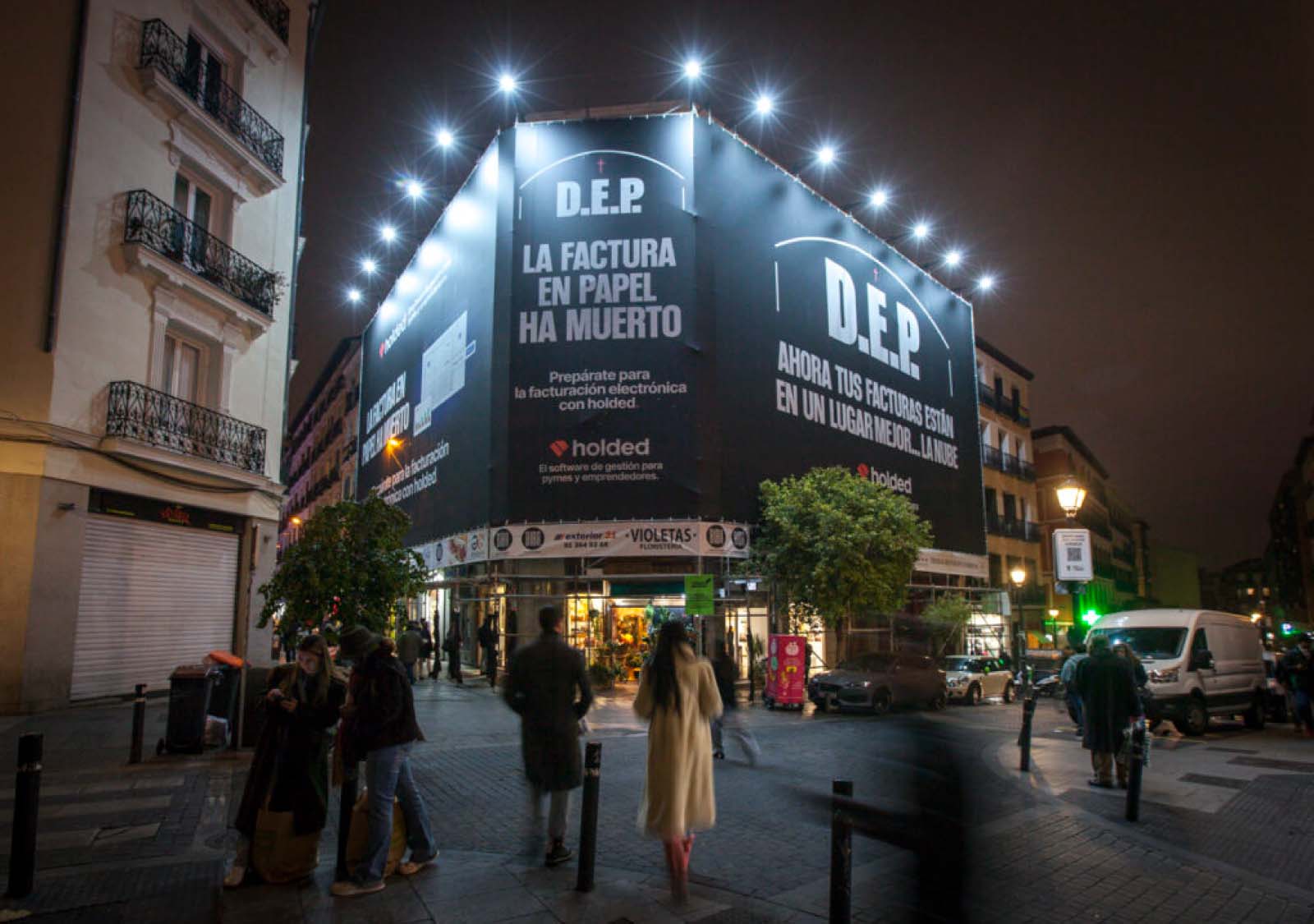 Anuncio publicitario en la fachada de un edificio donde se lee 'D.E.P.' (Descanse en Paz) y el logo de Holded, anunciando el fin de las facturas en papel y promoviendo su software de facturación electrónica.