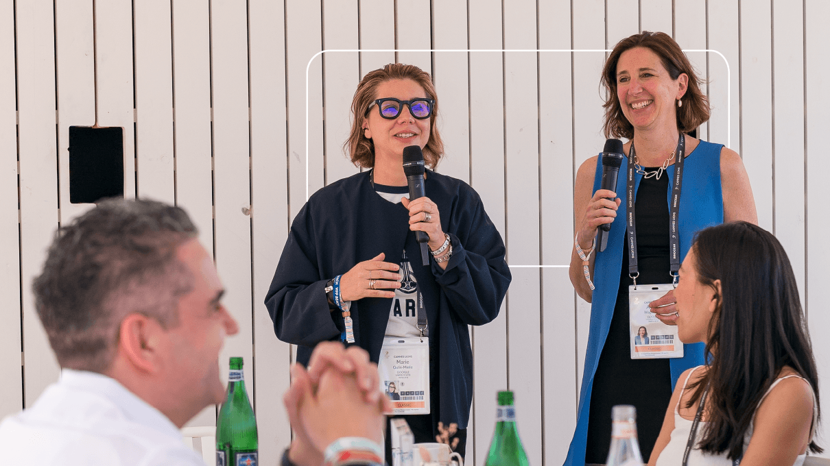 As VPs do Google, Marie Gulin-Merle e Alison Wagonfeld, falam em microfones para uma plateia de espectadores sentados à mesa com garrafas de água com gás e arranjos de flores. Todos sorriem.