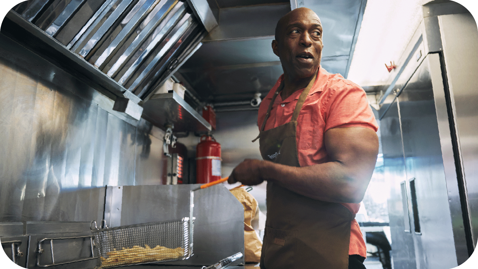 Jamie Boatright, wearing a brown apron over a red short-sleeved shirt, is pictured in his place of business, Treat Day Truck, holding a basket of french fries over a deep fryer.