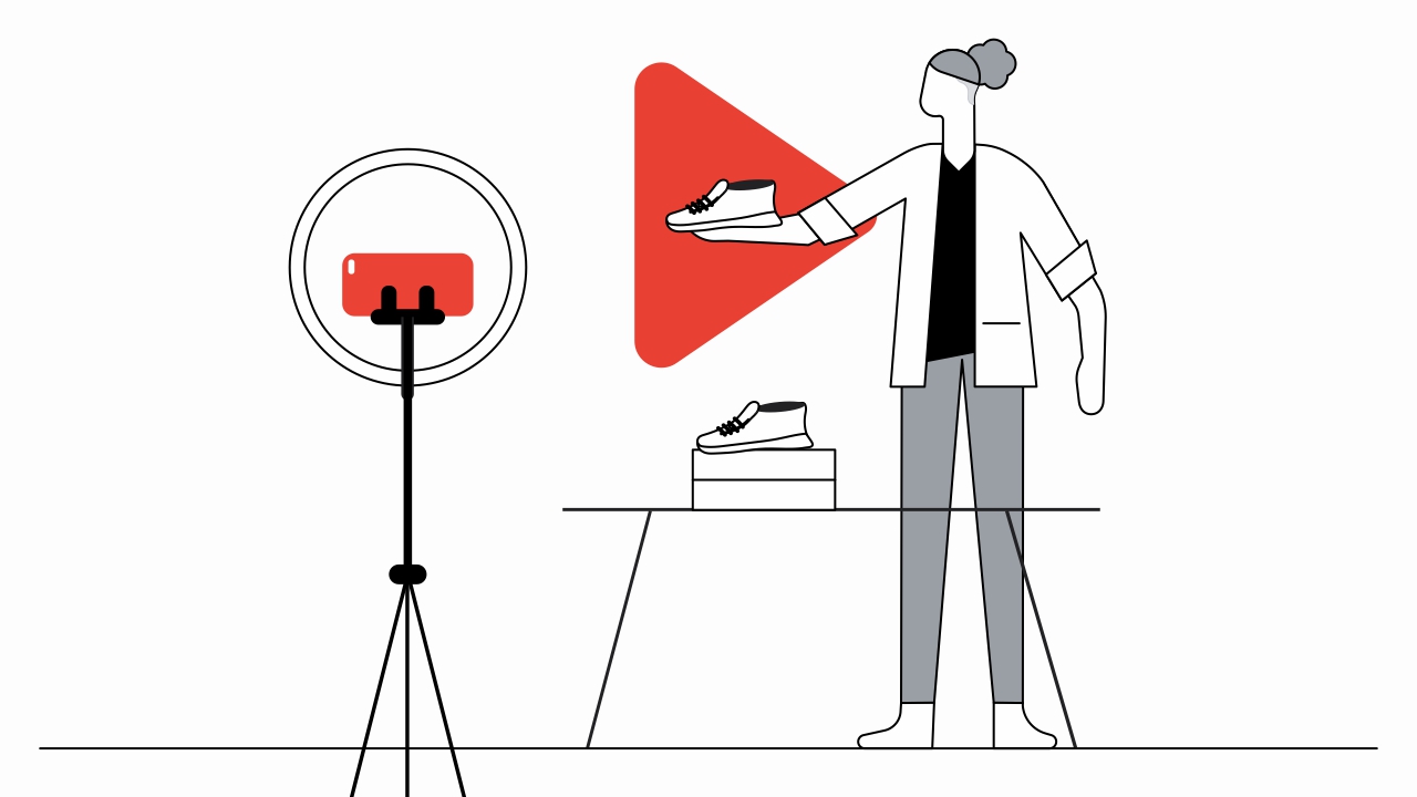 Una mujer con pelo enrulado y saco blanco muestra a la cámara un par de zapatos. Detrás de eso, el logo de YouTube en rojo. A la izquierda, un trípode con un teléfono móvil apuntando al zapato.