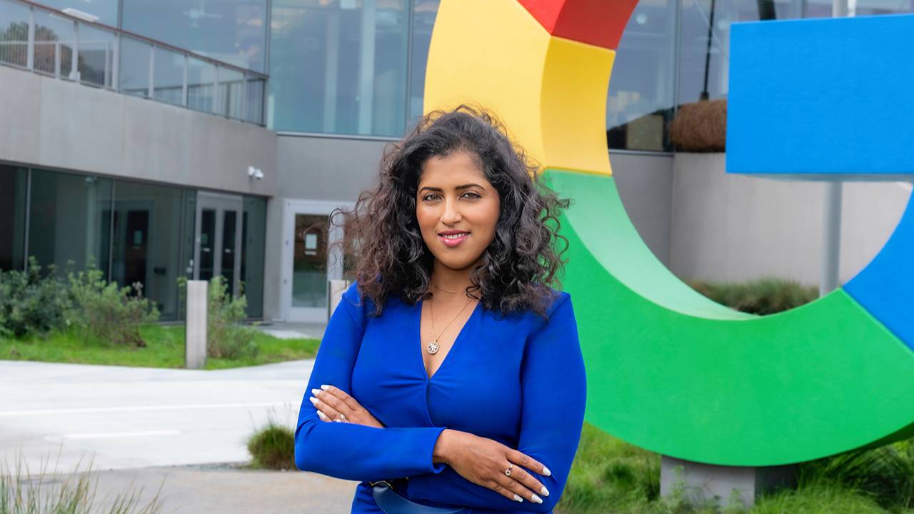 Vidhya Srinivasan, VP e General Manager di Google Ads presso Google, è in piedi fuori dall'ufficio aziendale di Mountain View, di fronte a una scultura gigante del logo G. Ha una carnagione media e i capelli castano scuro, ricci e lunghi e indossa un vest