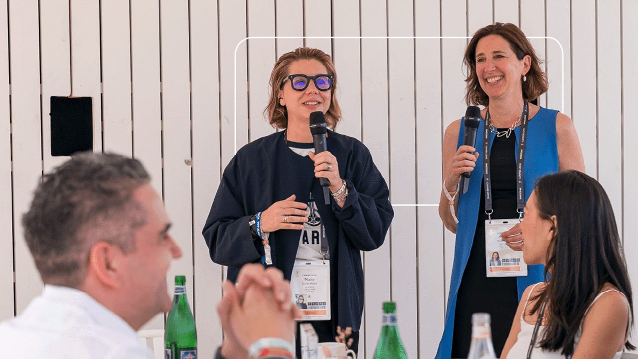 Les VP Google Marie Gulin-Merle et Alison Wagonfeld, un micro à la main, s'adressent à des participantes et des participants du Cannes Lions, assis à des tables sur lesquelles sont posées des bouteilles d'eau pétillante et des fleurs séchées. Tout le mond