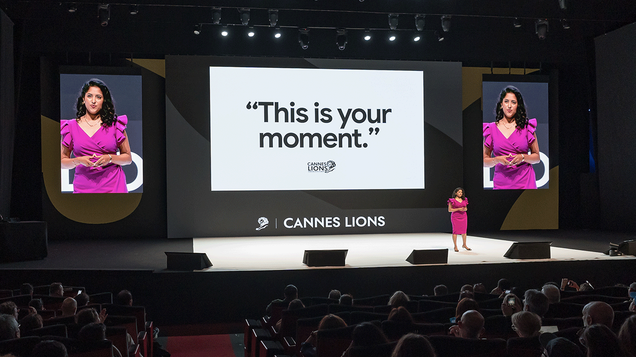 La Vicepresidenta y Directora General de Google Ads, Vidhya Srinivasan, pronuncia su discurso de apertura en el Palais Stage de Cannes. Con un vestido rosa intenso, se encuentra frente a una enorme pantalla en la que se lee "This is your moment".