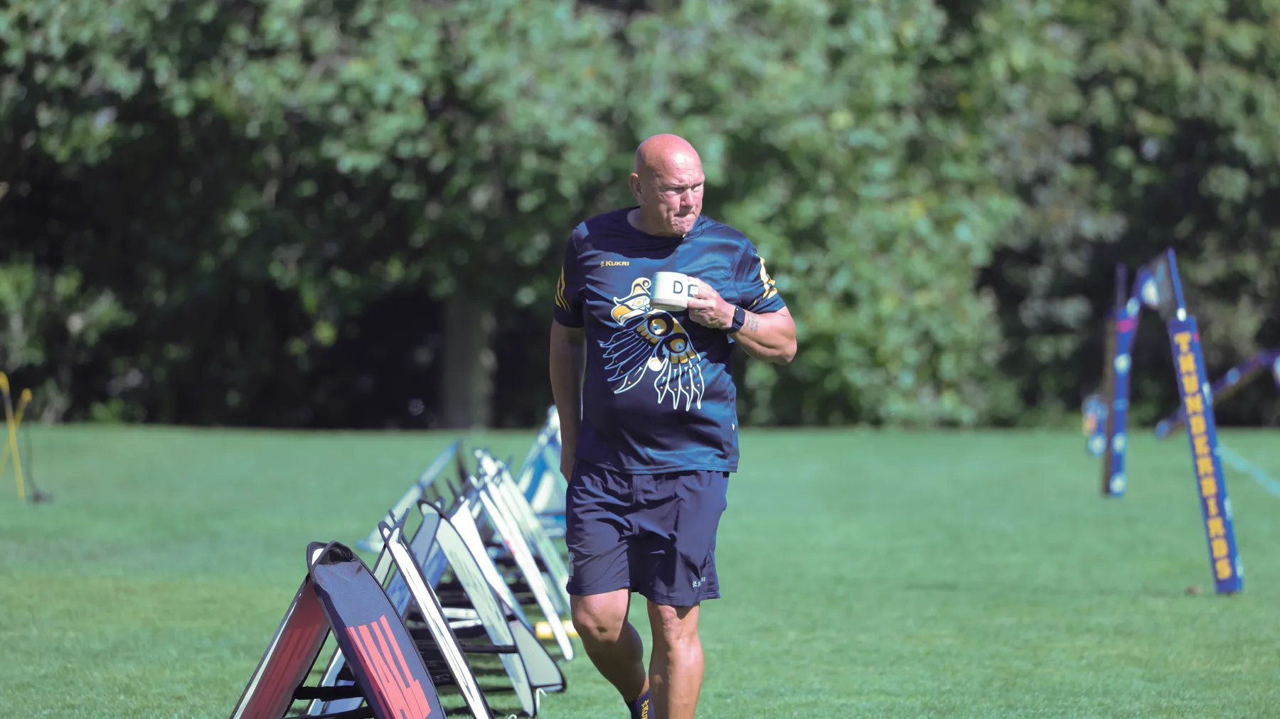A bald man wear a navy blue rugby jersey kit with an Indigenous-designed Thunderbird shirt faces the camera with his face turned slightly to the right. He has a cream-coloured mug that has a "D" on it and he is wearing a watch. The man is standing outside, on the sidelines of a green rugby field with trees in the background.