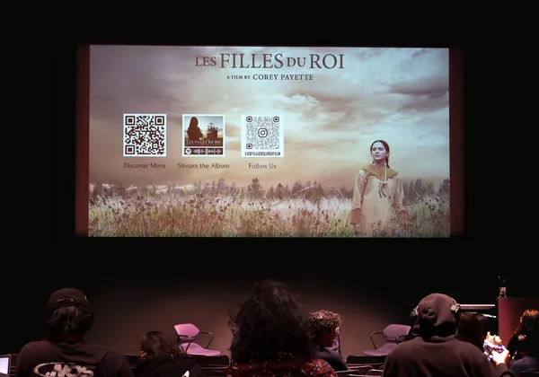 The backs of people's heads as they sit in a theatre looking at a large screen. The screen that has the words "Les Filles du Roi" and a photo of a young girl standing in a field, as well as two QR codes.