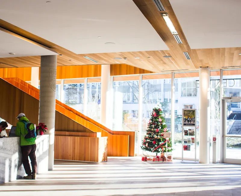 The image shows the inside of the UBC alumni centre, primarily featuring the bright wooden staircase leading upstairs and a Christmas tree is in front of the stairs.