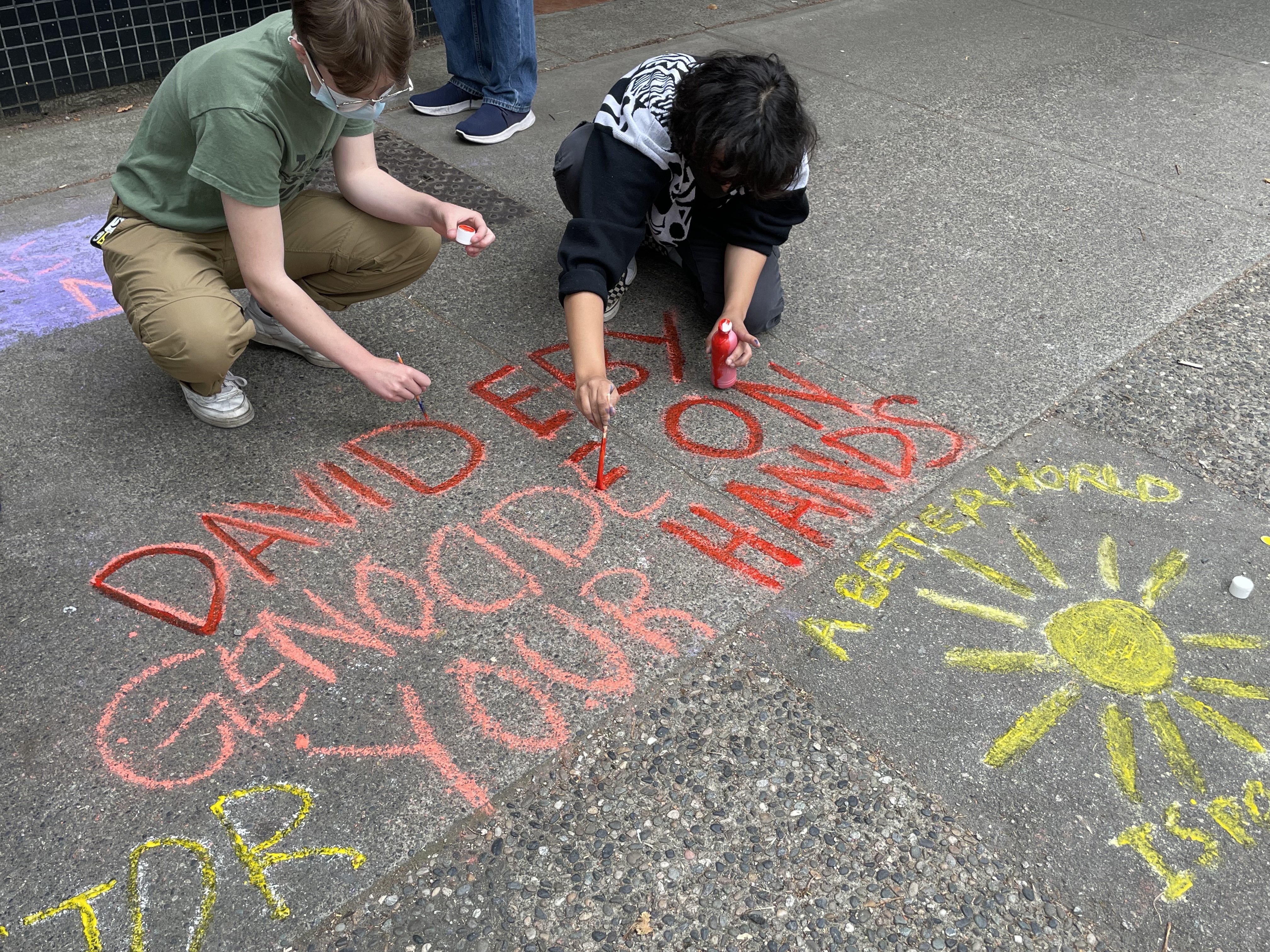 Two protestors paint over a chalk-written message on the sidewalk in front of Eby's office.