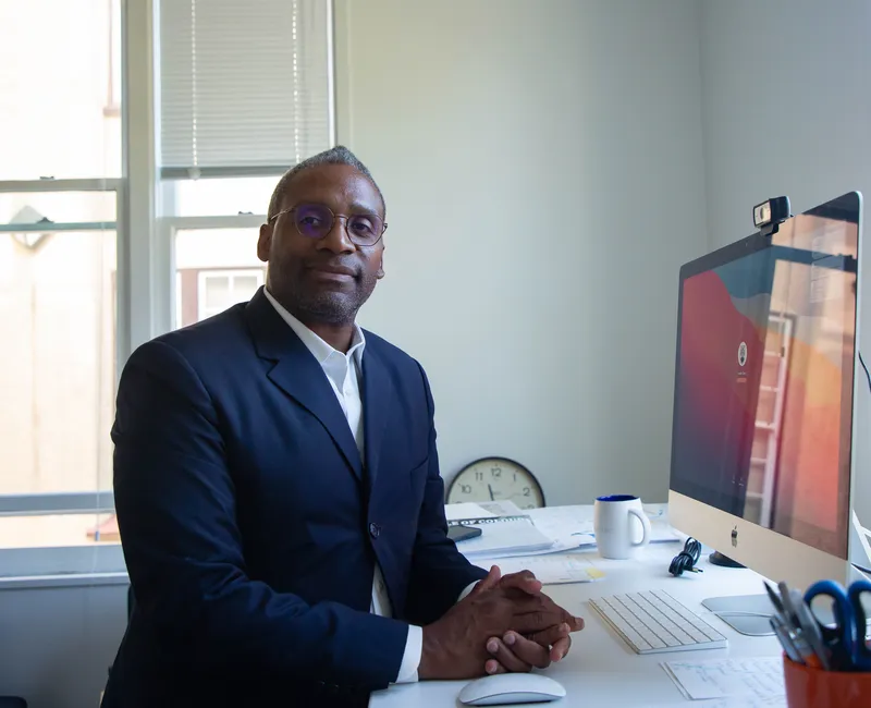 Aisnley Carry UBC seated at his office desk, surrounded by work materials, a computer, and office tools, indicating a professional workspace setting on campus.