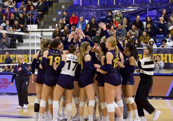 A photo of a women's volleyball team in navy blue jersey kits huddled on a court. Their hands are all in the raised in the middle ready to cheer. They stand on an indoor basketball court and there are fans in the stands behind them.