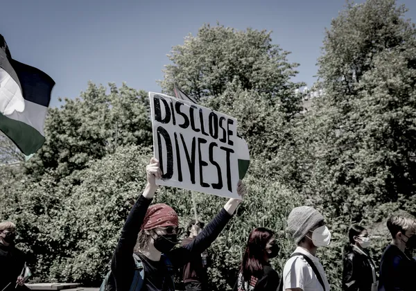 A close up photograph of a white sign at a protest, with the words "DISCLOSE" and "DIVEST" written in black and held up by a demonstrator.