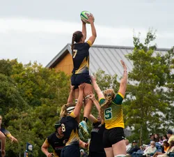 A rugby player wearing #7 catches the ball in the air, being lifted in a rugby lineout. She is held up alone, with the lineout not being contested.