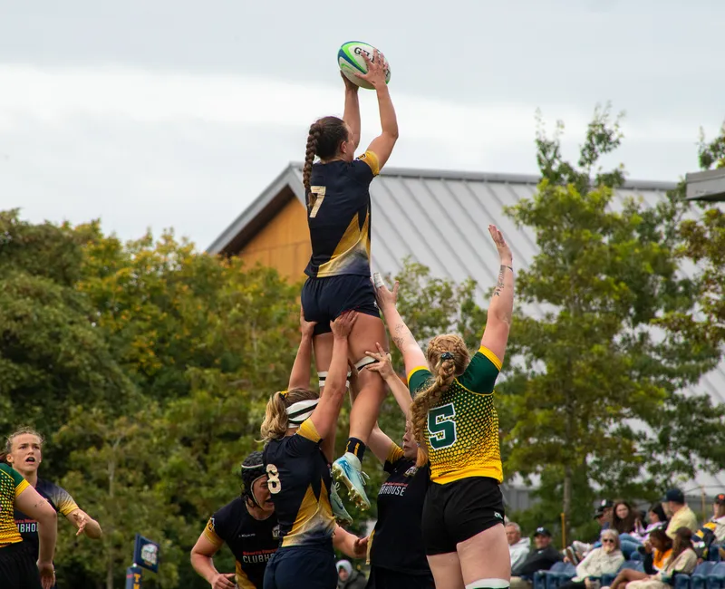 A rugby player wearing #7 catches the ball in the air, being lifted in a rugby lineout. She is held up alone, with the lineout not being contested.
