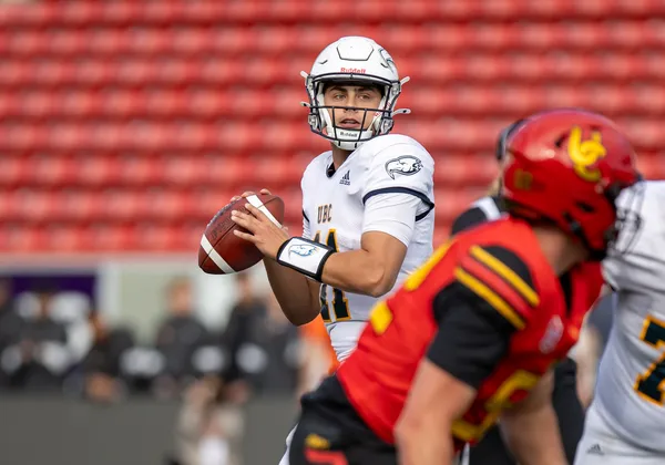 A football player in a white jersey kit prepares to throw the ball. A player in a red and black jersey is positioned in front of the other player, and slightly blurry. The background is empty red stands, but they are blurry.