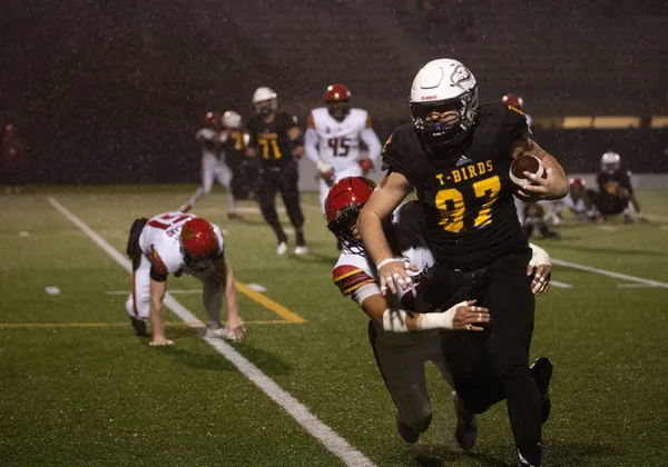 A football player in a navy blue jersey and a white helmet runs with the ball tucked in his left arm as another player in a red helmet and white jersey is attempting to wrap his arms around him. They are on a green football field at night, with visible raindrops in the background of the photo.