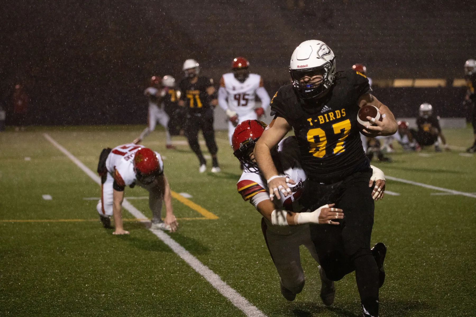 A football player in a navy blue jersey and a white helmet runs with the ball tucked in his left arm as another player in a red helmet and white jersey is attempting to wrap his arms around him. They are on a green football field at night, with visible raindrops in the background of the photo.