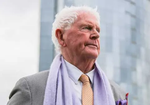 Peter Wall photographed wearing a grey suit, white collared shirt, orange tie and purple scarf in front of the One Wall Centre.