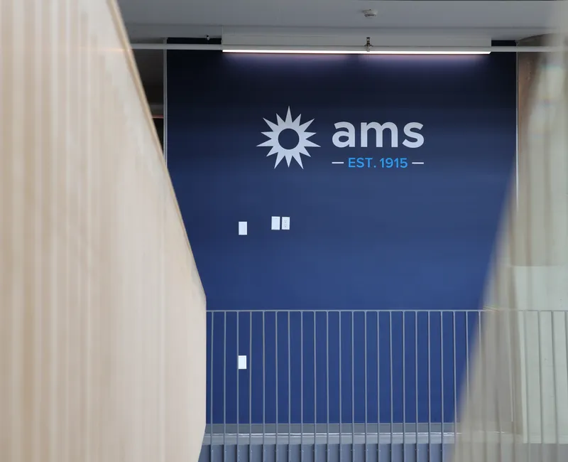 A photo inside the AMS Student Nest. The AMS logo is displayed on a blue wall, with railings in the foreground.