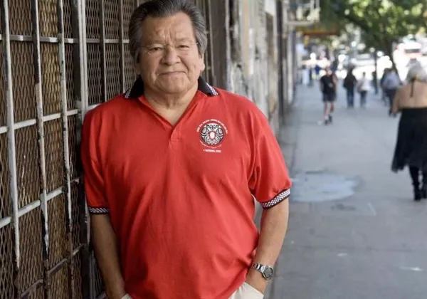 A photo of Bill Wilson smiling at the camera and leaning against a wall in an orange shirt.