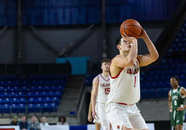 A man player in a white "Calgary" jersey aims for a free throw and the ball is in front of his face.