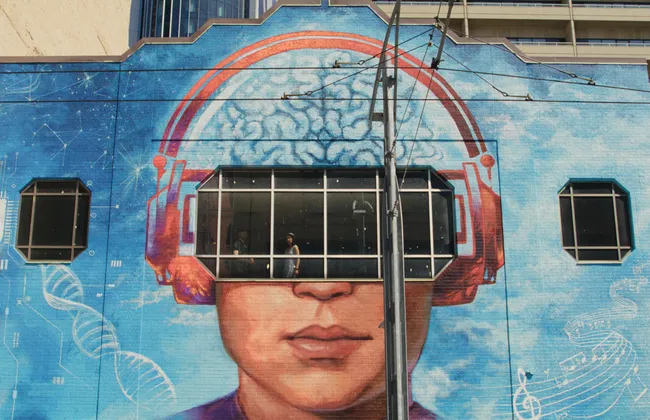 Shot from the exterior of a building with a brain-themed mural painted on the side, Radomski looks out a window.